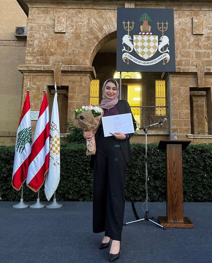 Hadeel holding flowers and her award certificate in front of the AUB crest