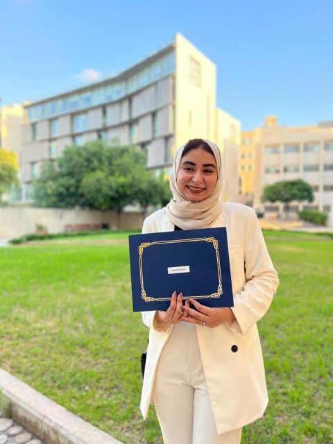 Hadeel holding a navy graduation folder on campus