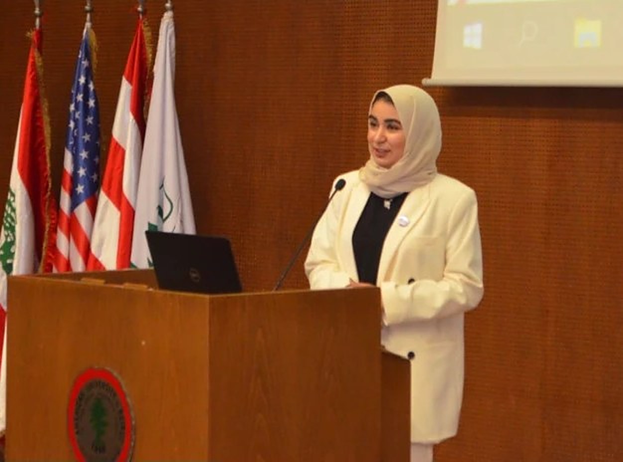 Hadeel speaking at a podium in front of Lebanese, American and university flags
